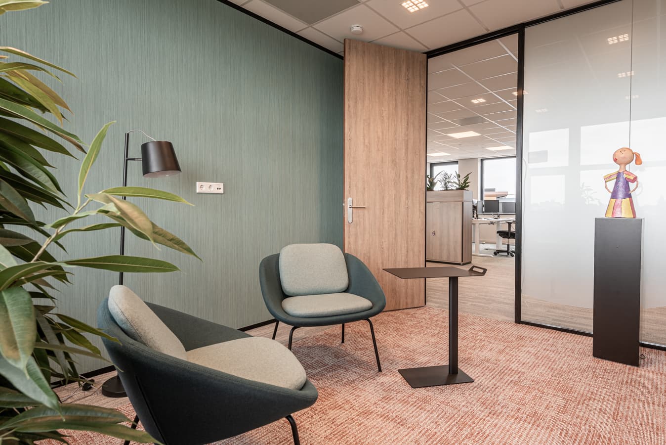 Informele overlegplek met twee fauteuils, een kleine tafel en een glazen scheidingswand in een kantoor.