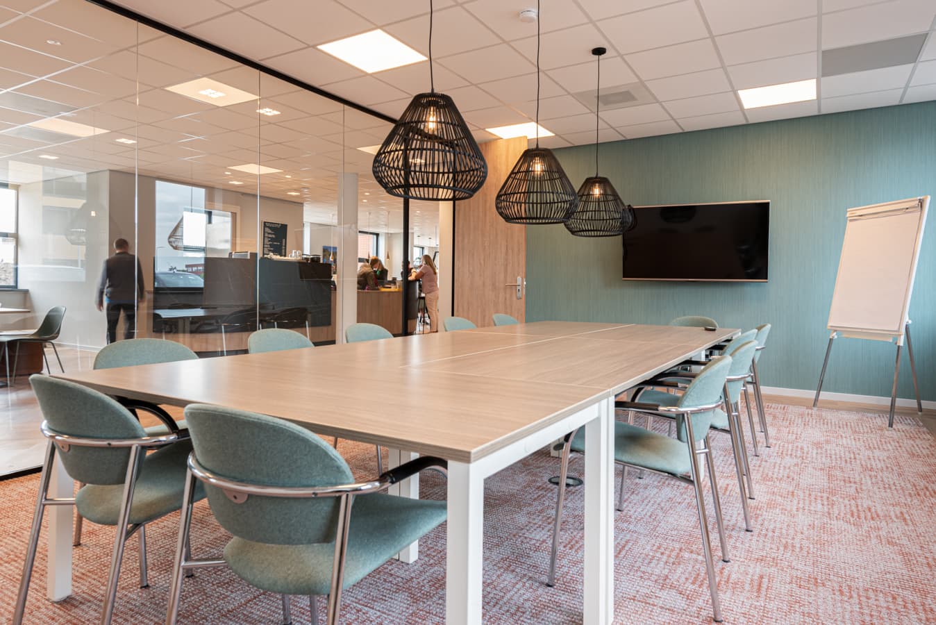 Vergaderruimte met grote houten tafel, groene stoelen, beeldscherm en flip-over