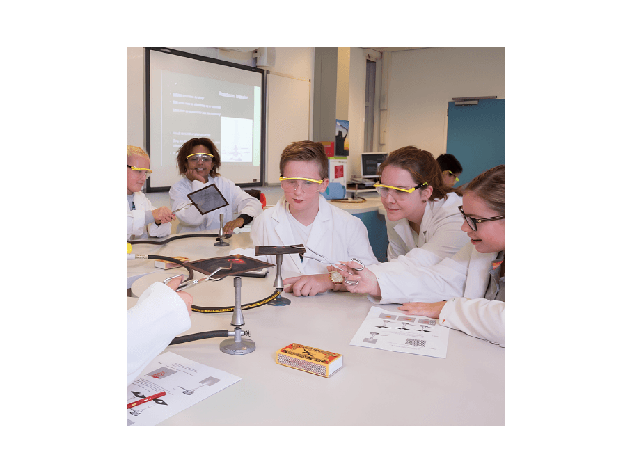 Leerlingen in labjassen en veiligheidsbrillen voeren een practicum uit aan een werktafel.