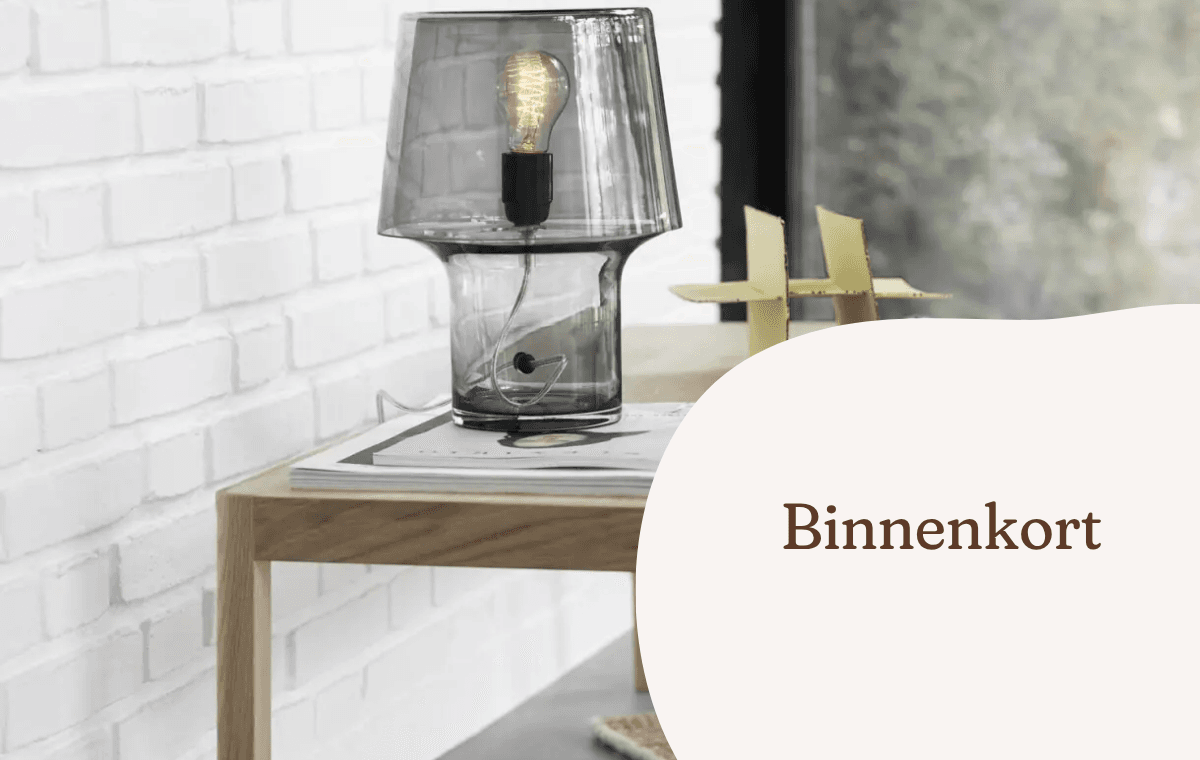 Glazen tafellamp op een houten bijzettafel naast een witte bakstenen muur.