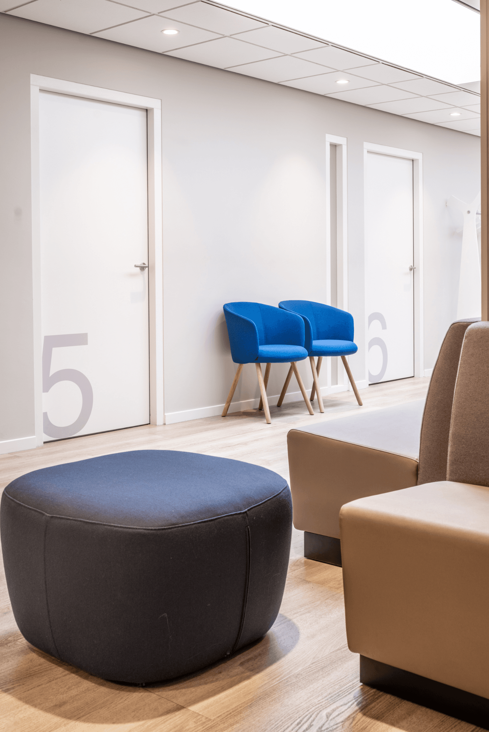 Wachtruimte met een taupekleurige zitbank, blauwe stoelen en een ronde poef