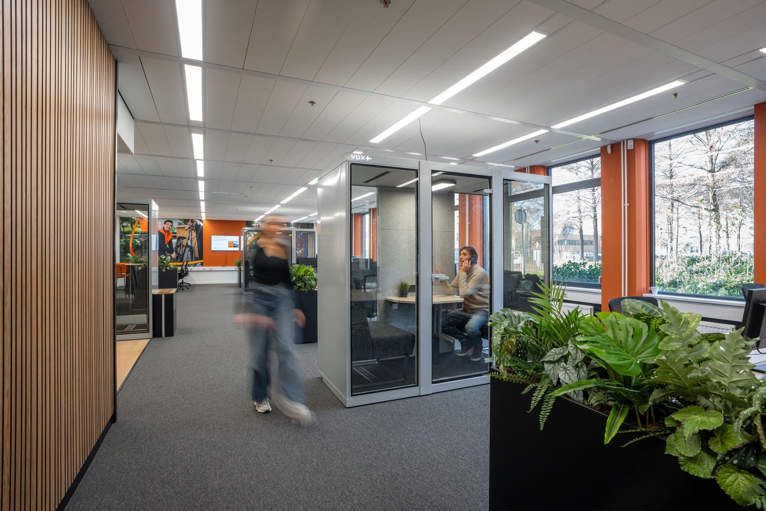 Medewerker loopt langs een glazen vergaderruimte in een kantoor met planten