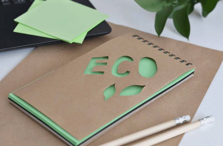 Notitieboek met groene accenten op een bureau naast potloden, laptop en een plant.