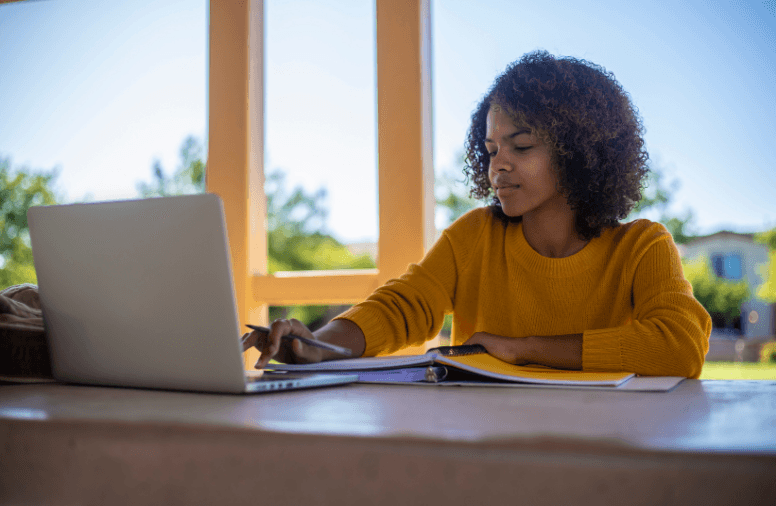 Vrouw in een okergele trui zit achter haar computer met een pen in haar hand en een schrijfblok ander haar arm